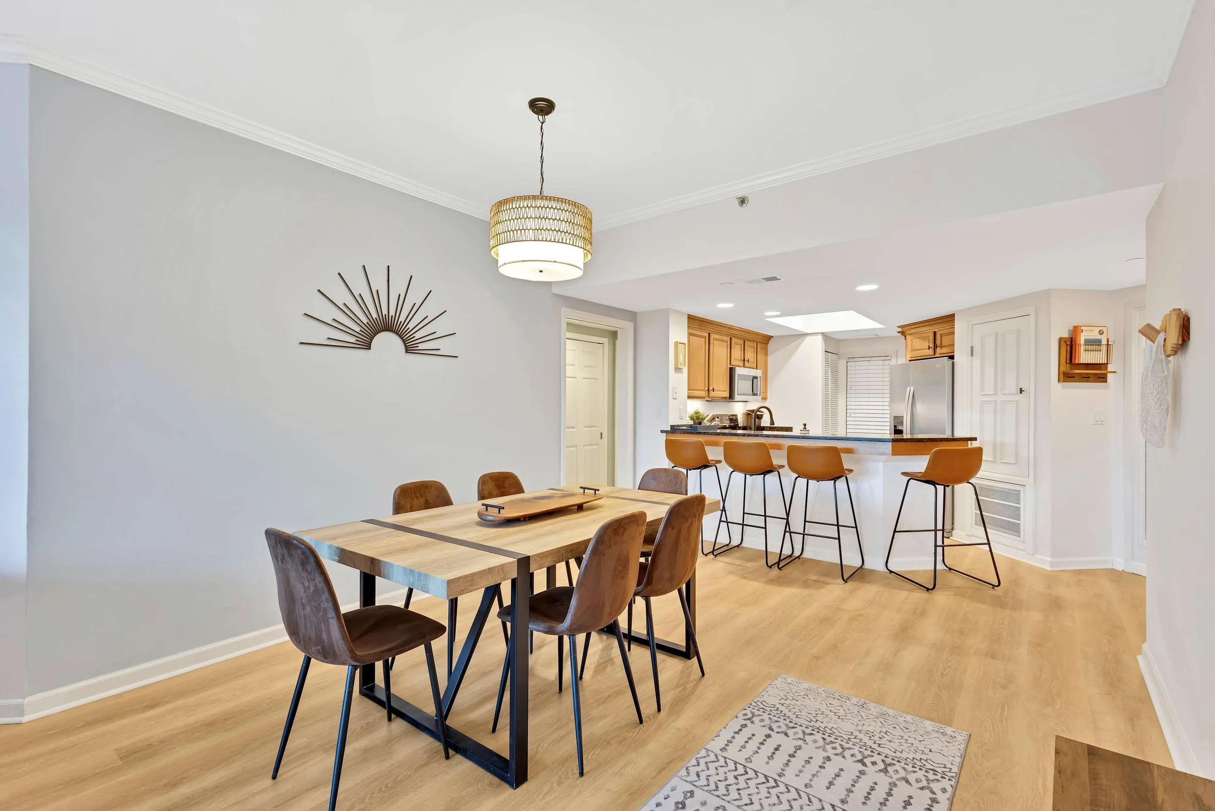 Modern dining and kitchen area with wooden table, six brown chairs, bar counter with high stools, pendant light, and wall decoration.