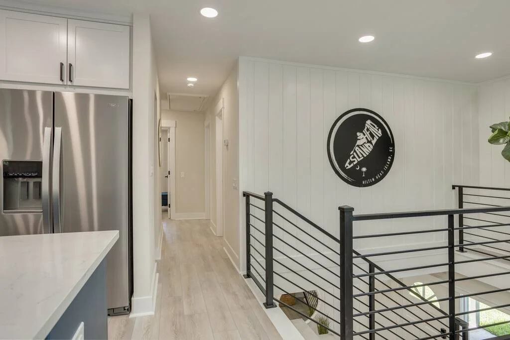 Modern interior hallway with stainless steel refrigerator, white cabinets, black metal railing, and a round wall decoration featuring a surfboard design.