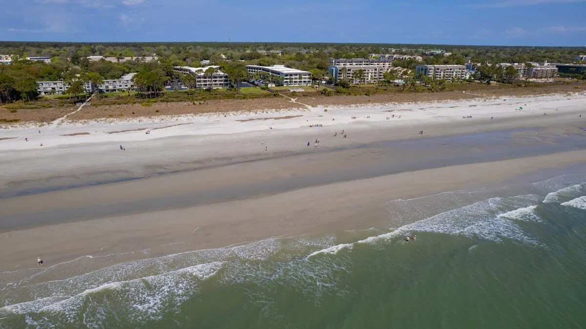 Aerial view of a sandy beach with gentle waves and a distant row of buildings surrounded by trees, under a clear blue sky.
