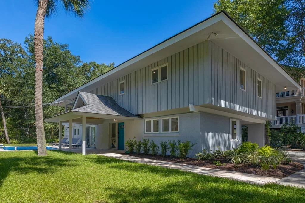 Modern two-story house with gray siding, large windows, and a front porch. The yard features a well-maintained lawn and a tall palm tree. In the background, there's a clear blue sky and surrounding greenery.