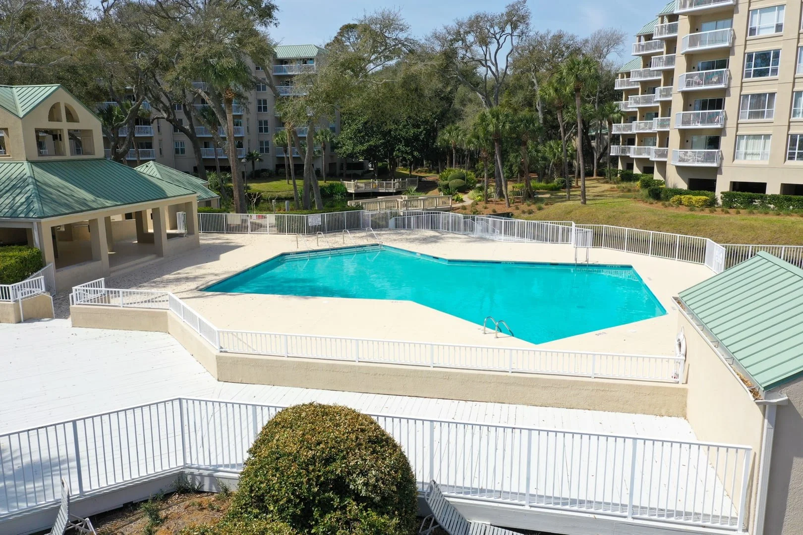 Residential swimming pool area with white fencing, surrounded by buildings and trees.