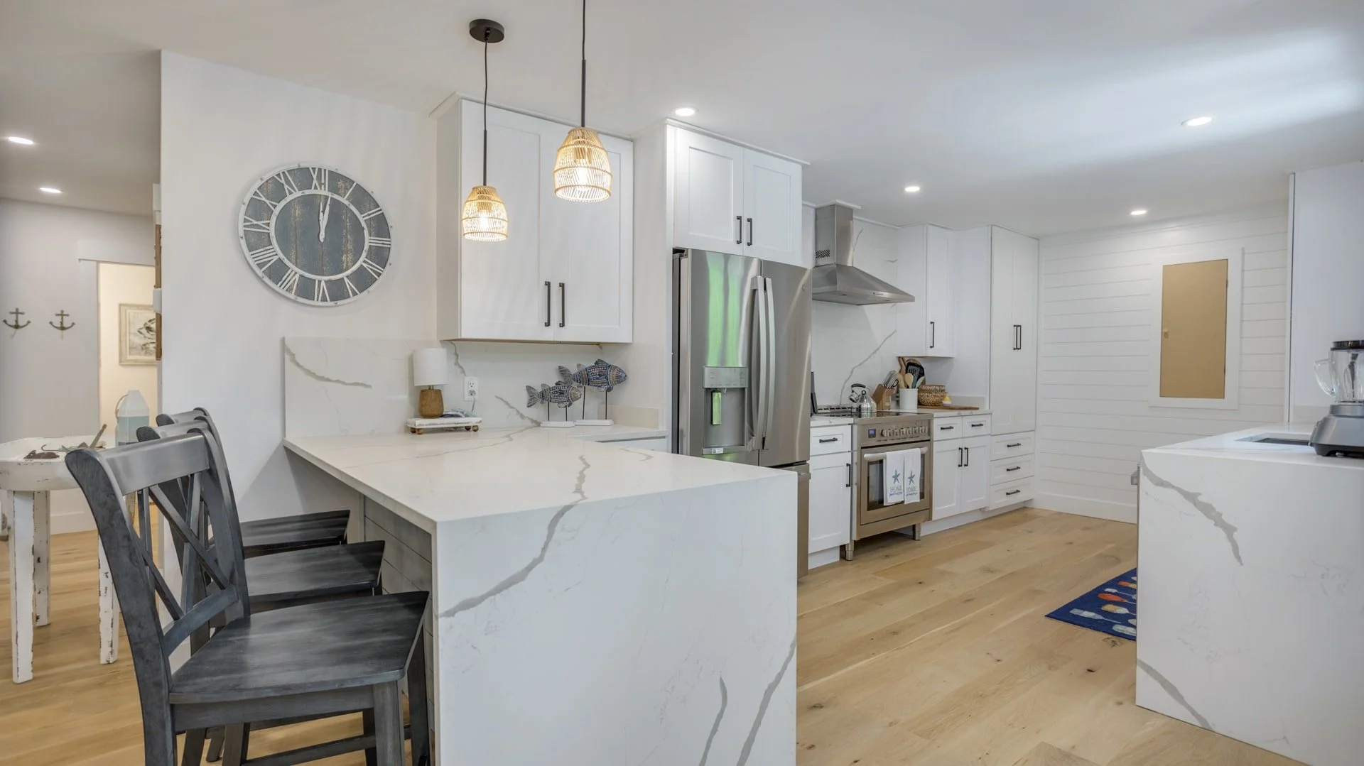 Modern kitchen interior with white cabinets, stainless steel appliances, and a marble countertop island. A large wall clock hangs above the counter. Pendant lights are suspended above the island, with black bar stools underneath. The room has a light