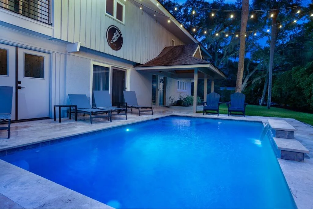 Outdoor pool area with lounge chairs and string lights, adjacent to a modern house at dusk.