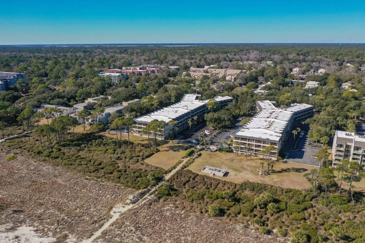 Aerial view of a residential complex with buildings surrounded by greenery and trees, set in a forested area near a sandy landscape under a clear blue sky.