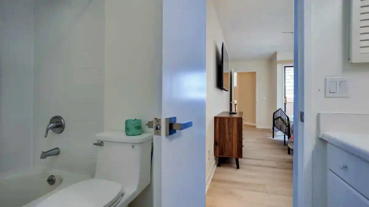 View from a bathroom with a bathtub, toilet, and green toilet paper, looking into a bedroom with wooden flooring, a dresser, wall-mounted TV, and a bed.