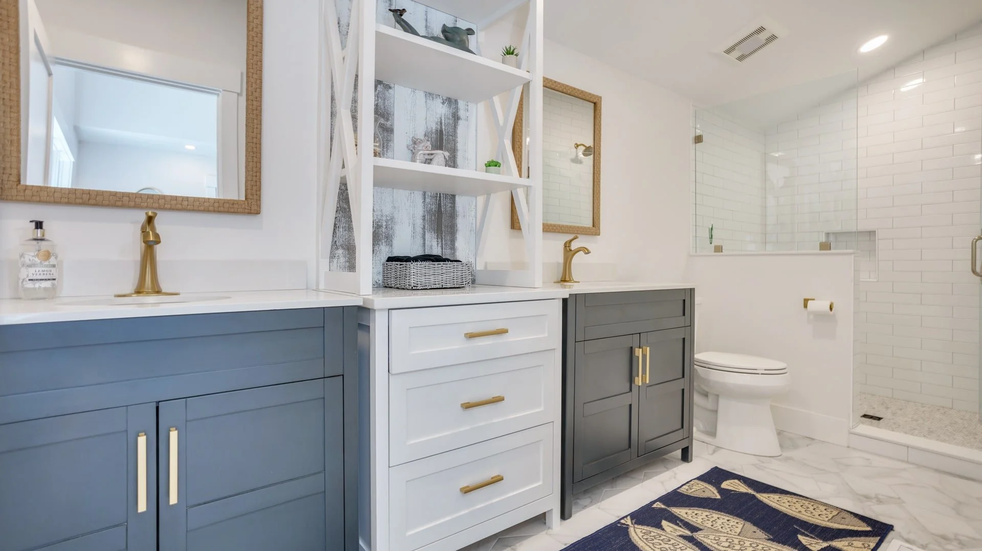 Modern bathroom with blue and white cabinetry, a white countertop, gold fixtures, a toilet, a walk-in shower with glass doors, a woven mirror, and decorative shelving.