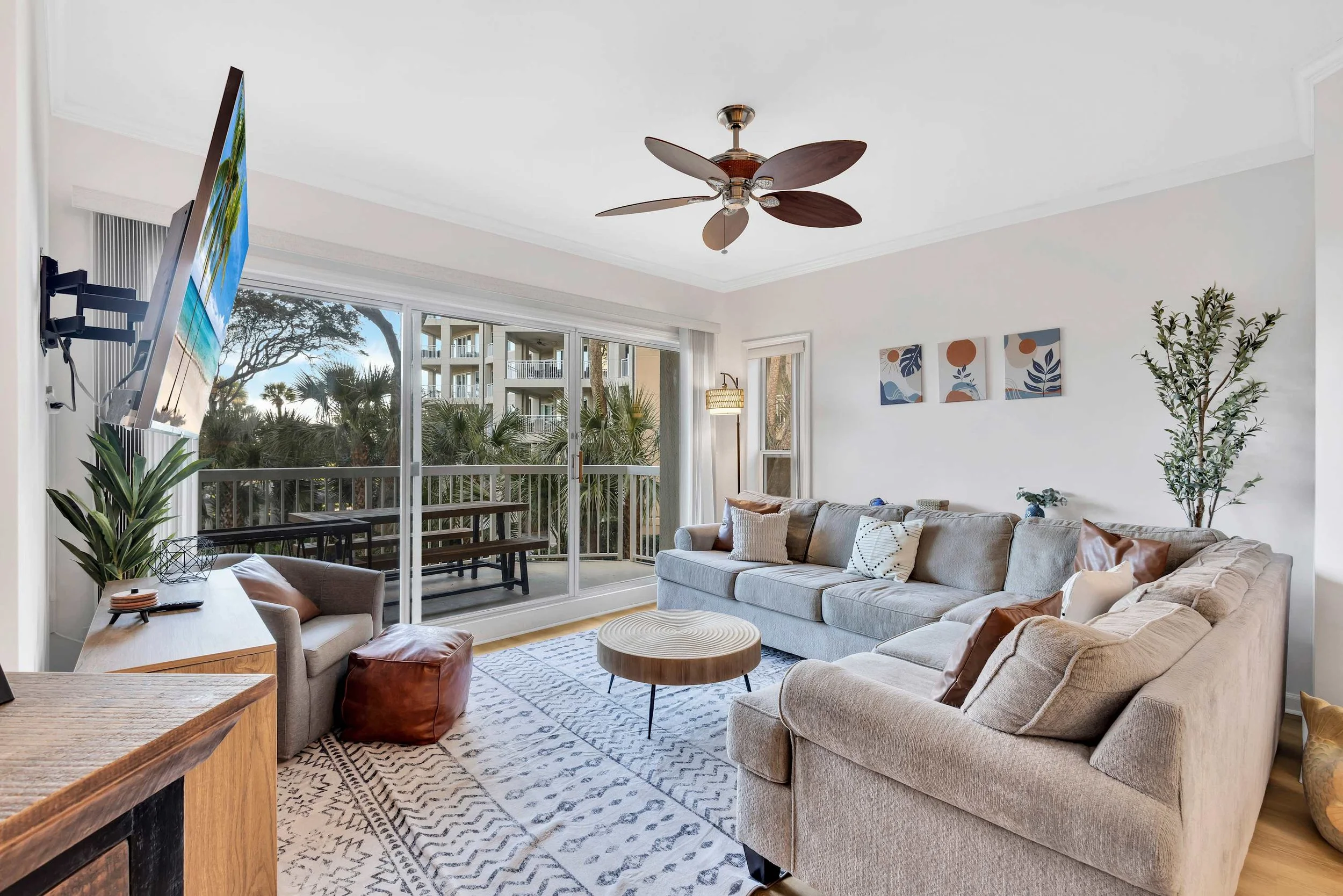Modern living room with beige sectional sofa, round wooden coffee table, wall-mounted TV, ceiling fan, decorative plants, and sliding glass doors leading to a balcony with outdoor seating.