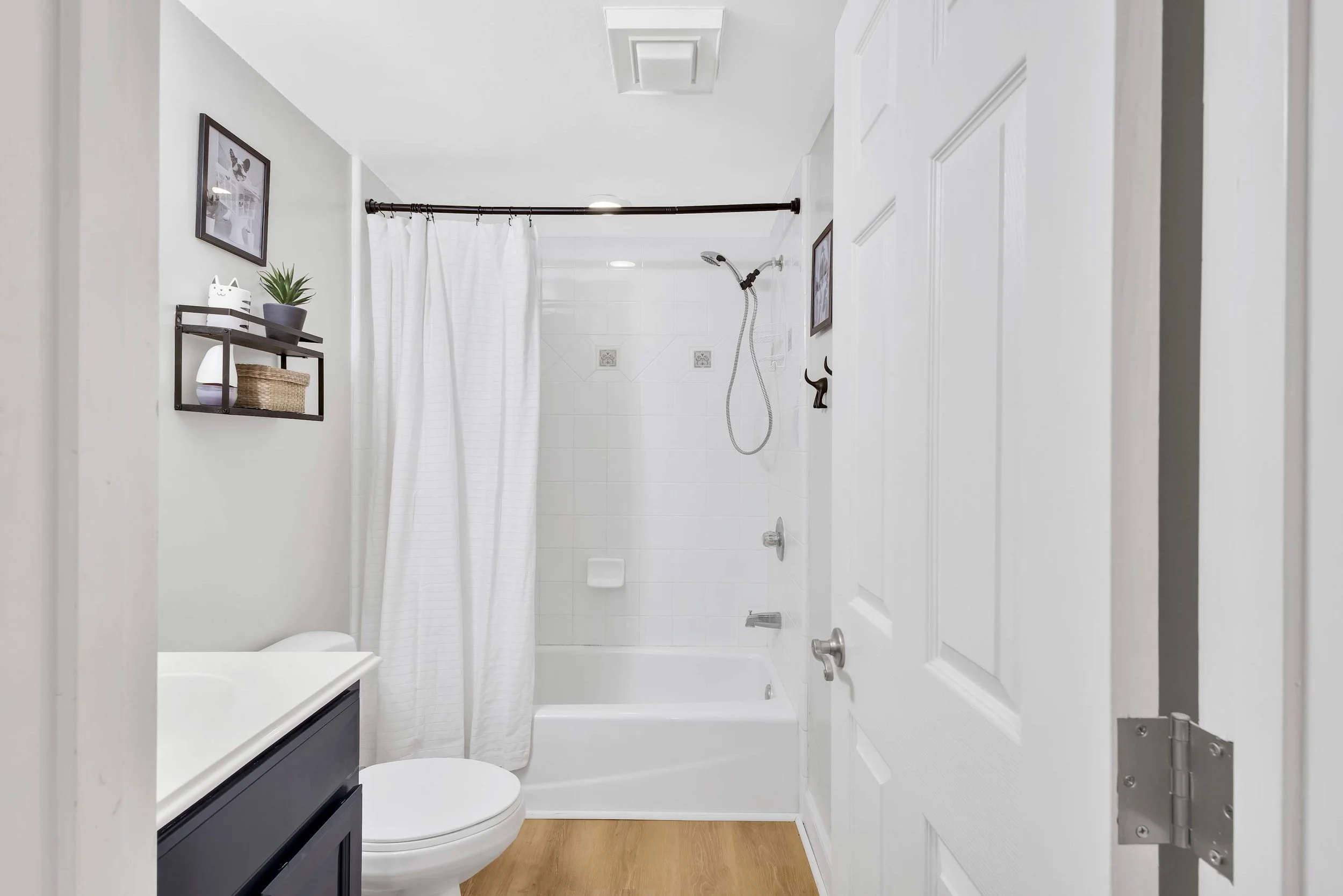 Modern bathroom with white shower curtain, bathtub, toilet, and sink. Wall shelves hold a plant and decor items. Wooden floor adds warmth.