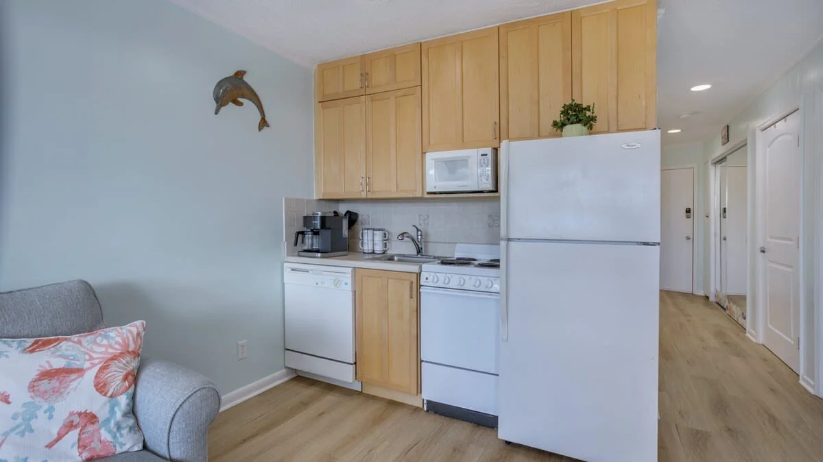 Small kitchen with wooden cabinets, white appliances including a refrigerator, microwave, dishwasher, and stove, and a dolphin wall decoration. A coffee maker is on the counter, and a plant is placed on top of the fridge. The wall is painted light bl