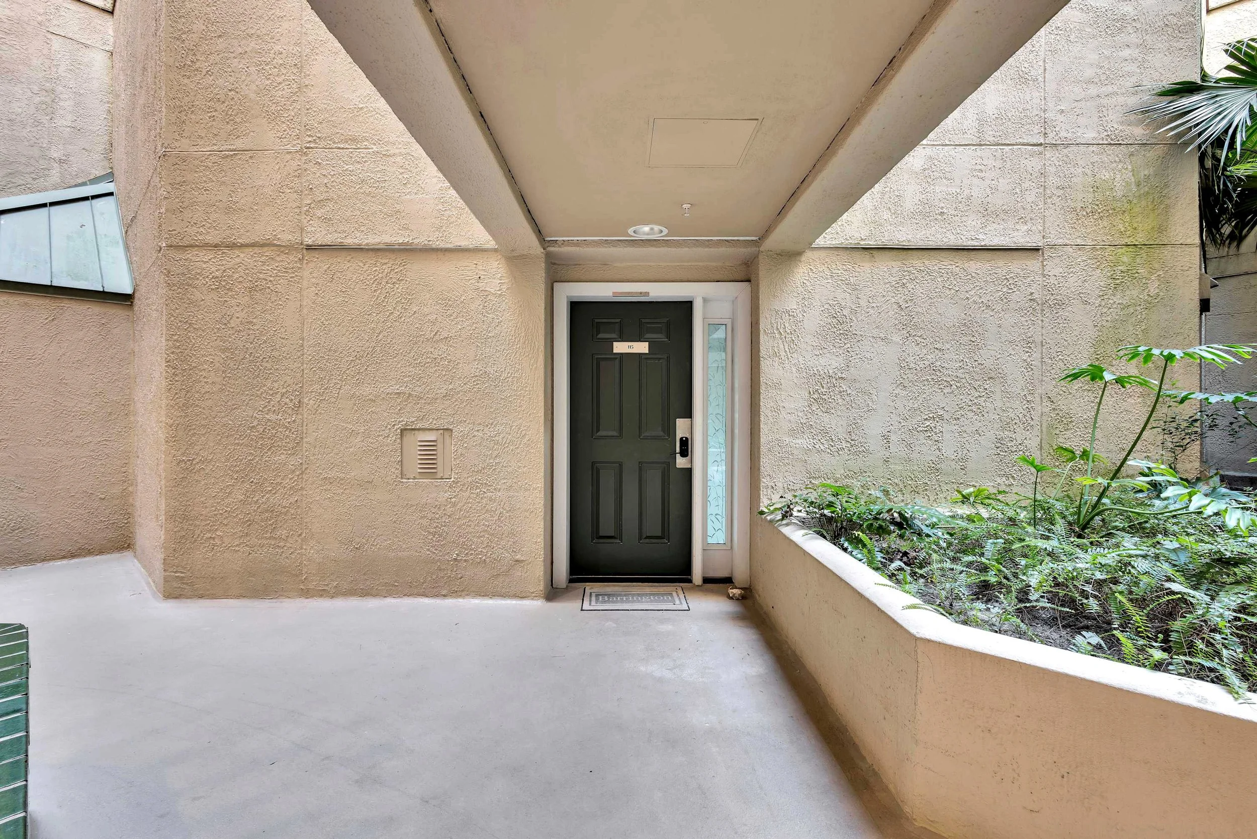 Exterior view of an apartment entrance with a green door, beige walls, and a side planter with green plants. The area has an overhead covering and concrete flooring.