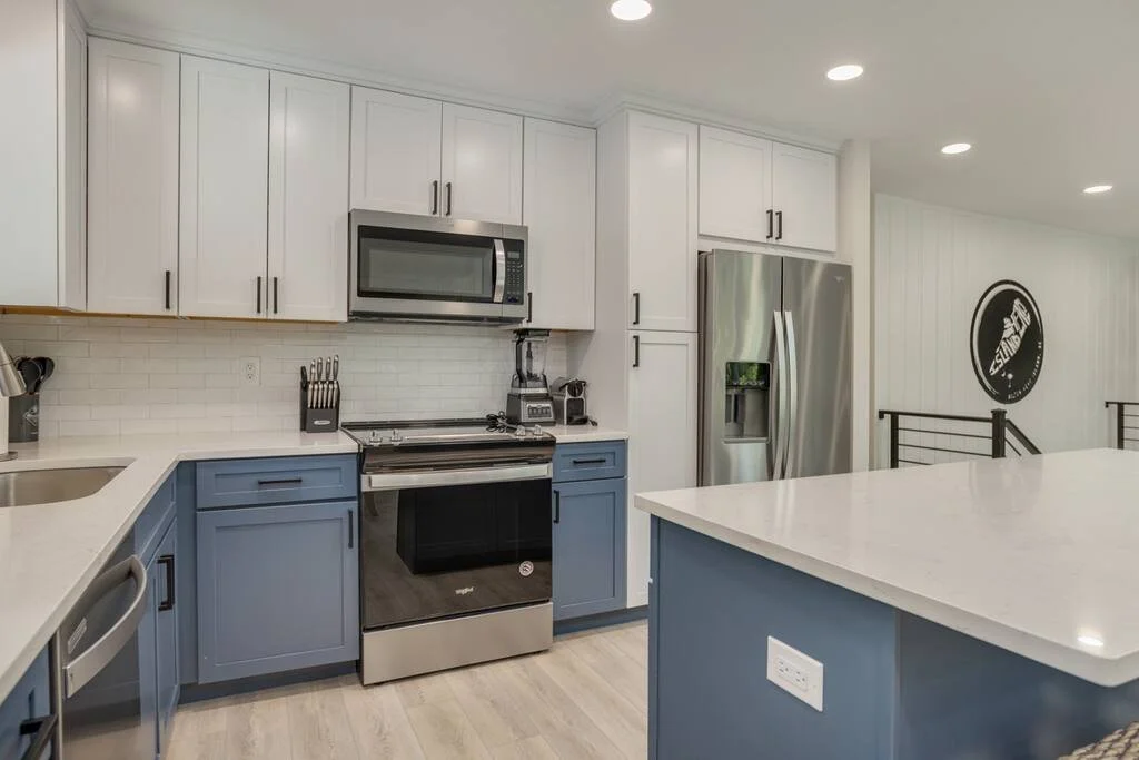 Modern kitchen with stainless steel appliances, including a refrigerator, oven, and microwave, white cabinetry, blue lower cabinets, a large kitchen island, and a light wood floor.