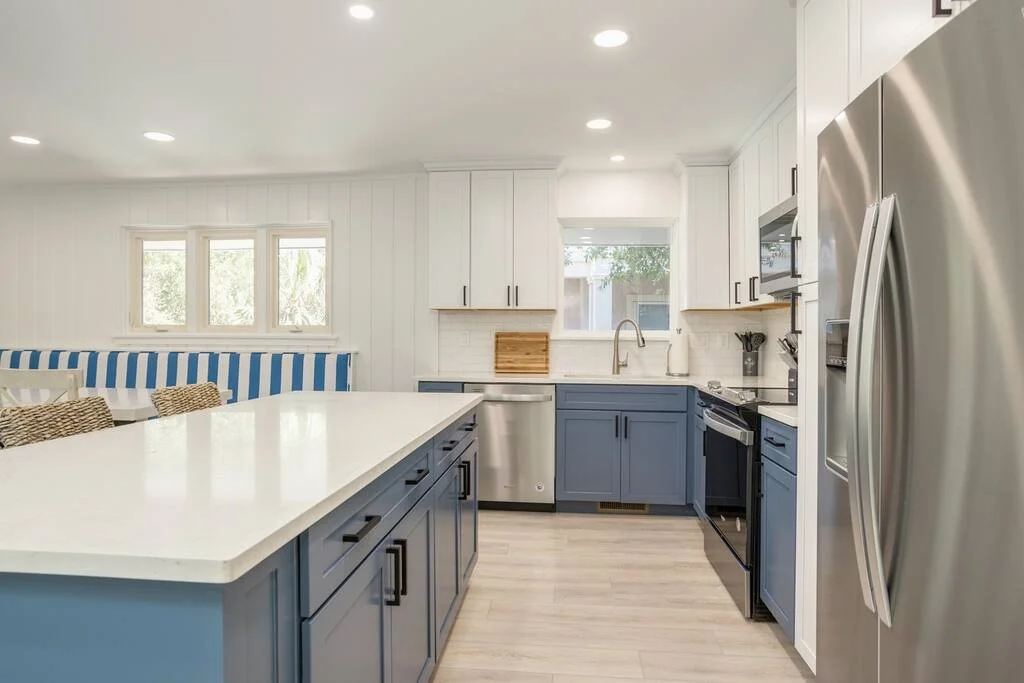 Modern kitchen with white and blue cabinets, stainless steel appliances, and a large island. The room is well-lit with recessed lighting and features a window view.