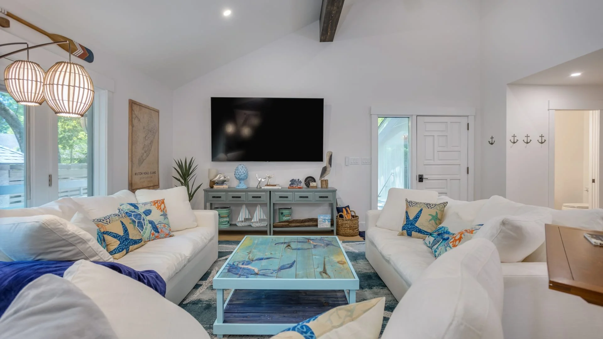 Coastal-themed living room with white sofas, colorful pillows, a large wall-mounted TV, decorative items, and a wooden coffee table.