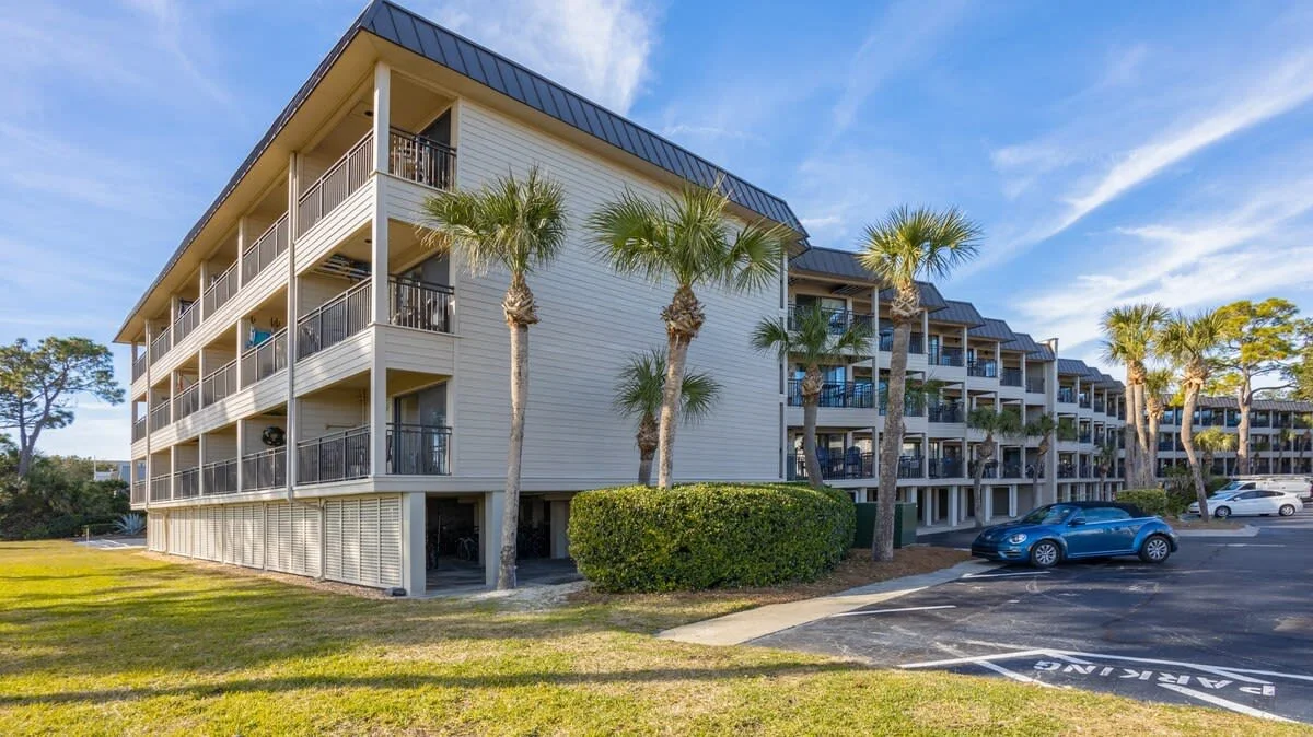 Three-story beachfront condominium with balconies, surrounded by palm trees and a parking lot with parked cars.