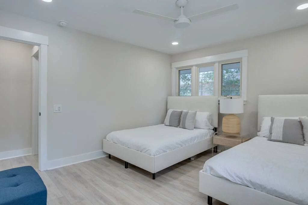 Bedroom with two white beds, a beige nightstand with a lamp, hardwood floor, ceiling fan, and a small window.