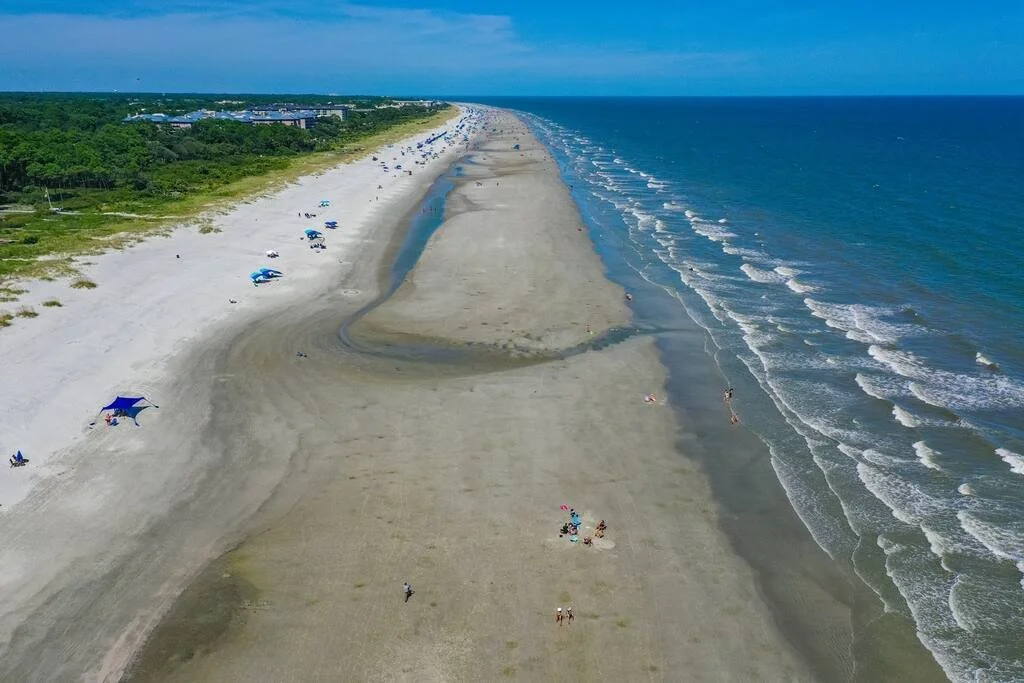 Aerial view of a wide sandy beach with people scattered along the shore, bordered by the ocean on the right and a line of trees and buildings on the left. The tide is low, revealing a large stretch of wet sand between the waves and the dry beach.