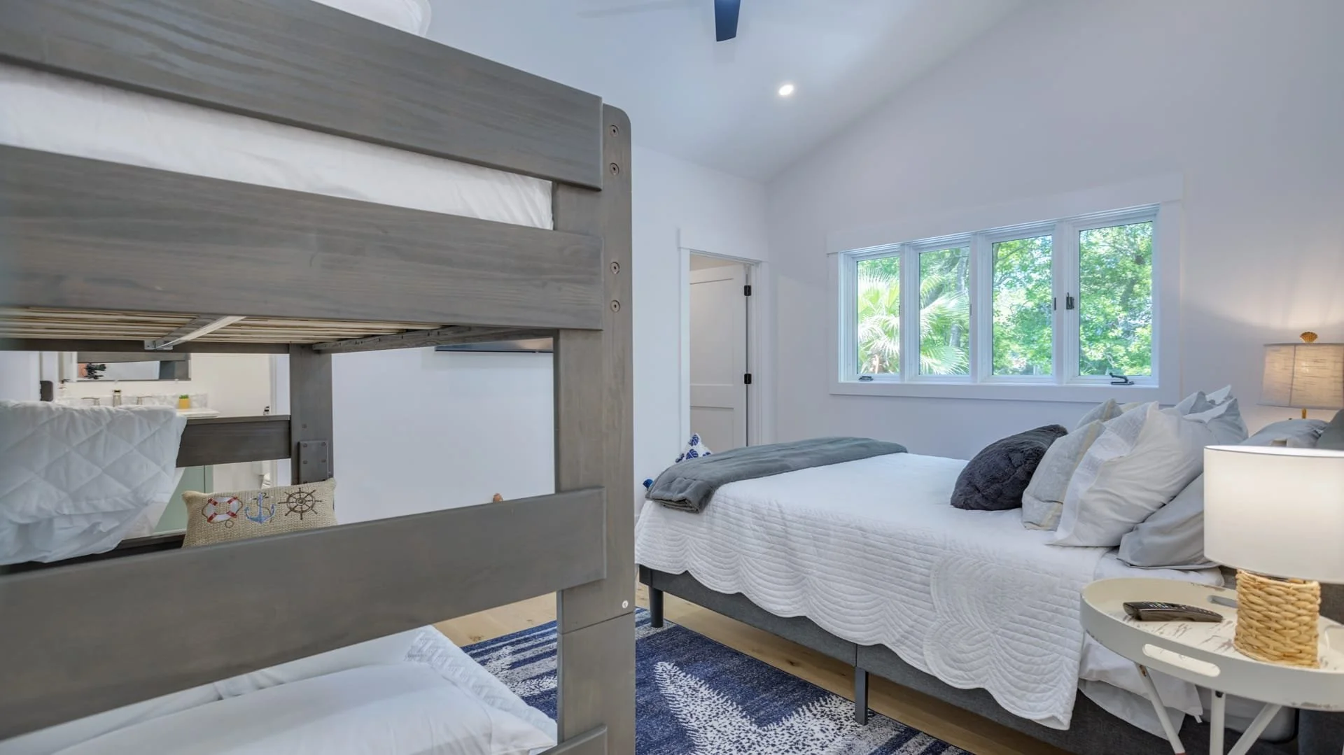 Bedroom with bunk beds and a double bed, white bedding, blue rug, window with green outdoor view, and bedside table with lamp and remote control.