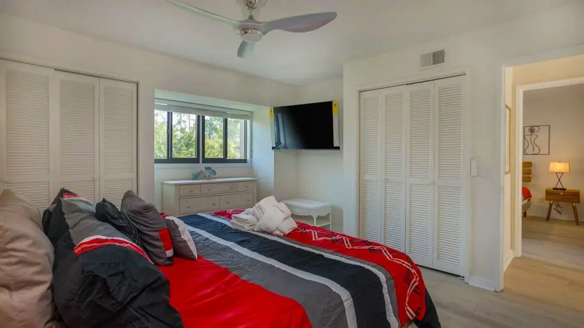 Modern bedroom with a colorful bedspread, ceiling fan, wall-mounted TV, large window, white dresser, and open closet doors.