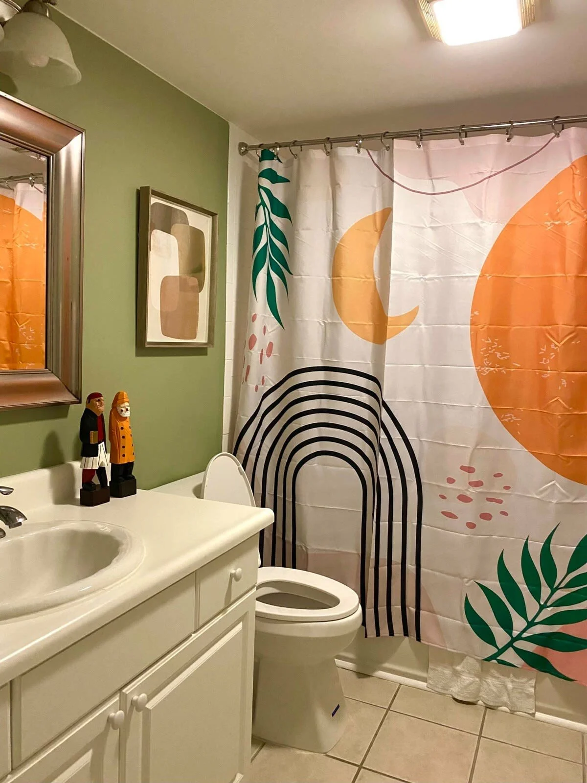 Bathroom with white vanity and toilet, featuring a colorful shower curtain with abstract shapes and plants. The walls are green with decorative art and two wooden figures on the counter.