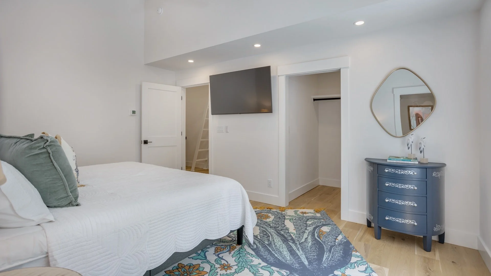 Modern bedroom with white bedding, decorative rug, blue dresser with mirror, wall-mounted TV, open closet, and wood flooring.