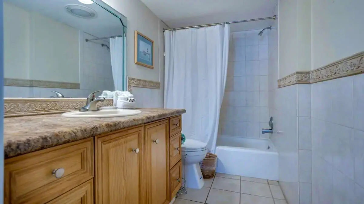 A modern bathroom with a wooden vanity, single sink, large mirror, and tiled walls. The bathtub has a white shower curtain. There is a toilet next to the bathtub and a decorative border along the walls.