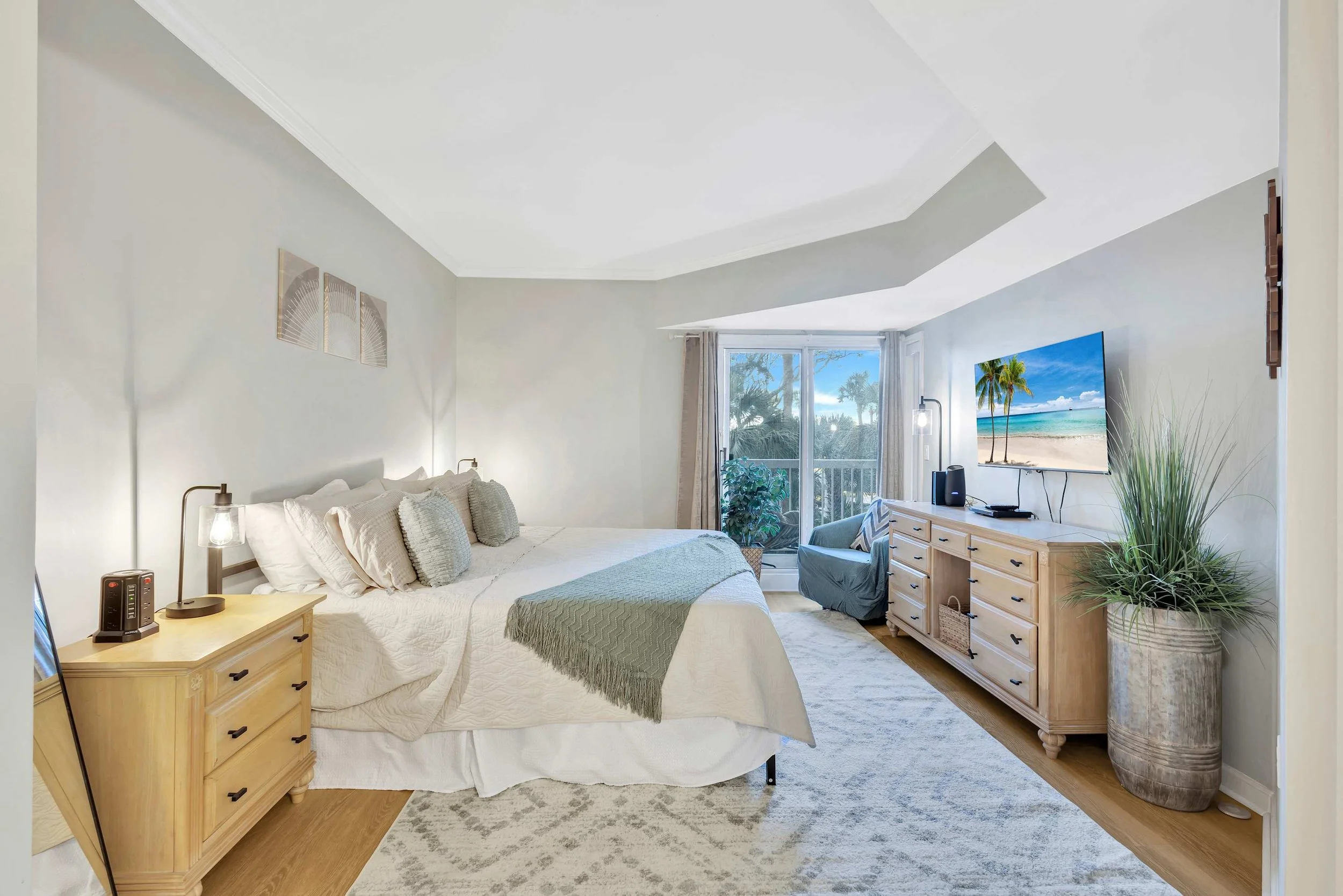 Cozy bedroom with king-sized bed, light wood furniture, and a large window with a view. The room features neutral tones, a TV displaying a beach scene, decorative pillows, and a potted plant.