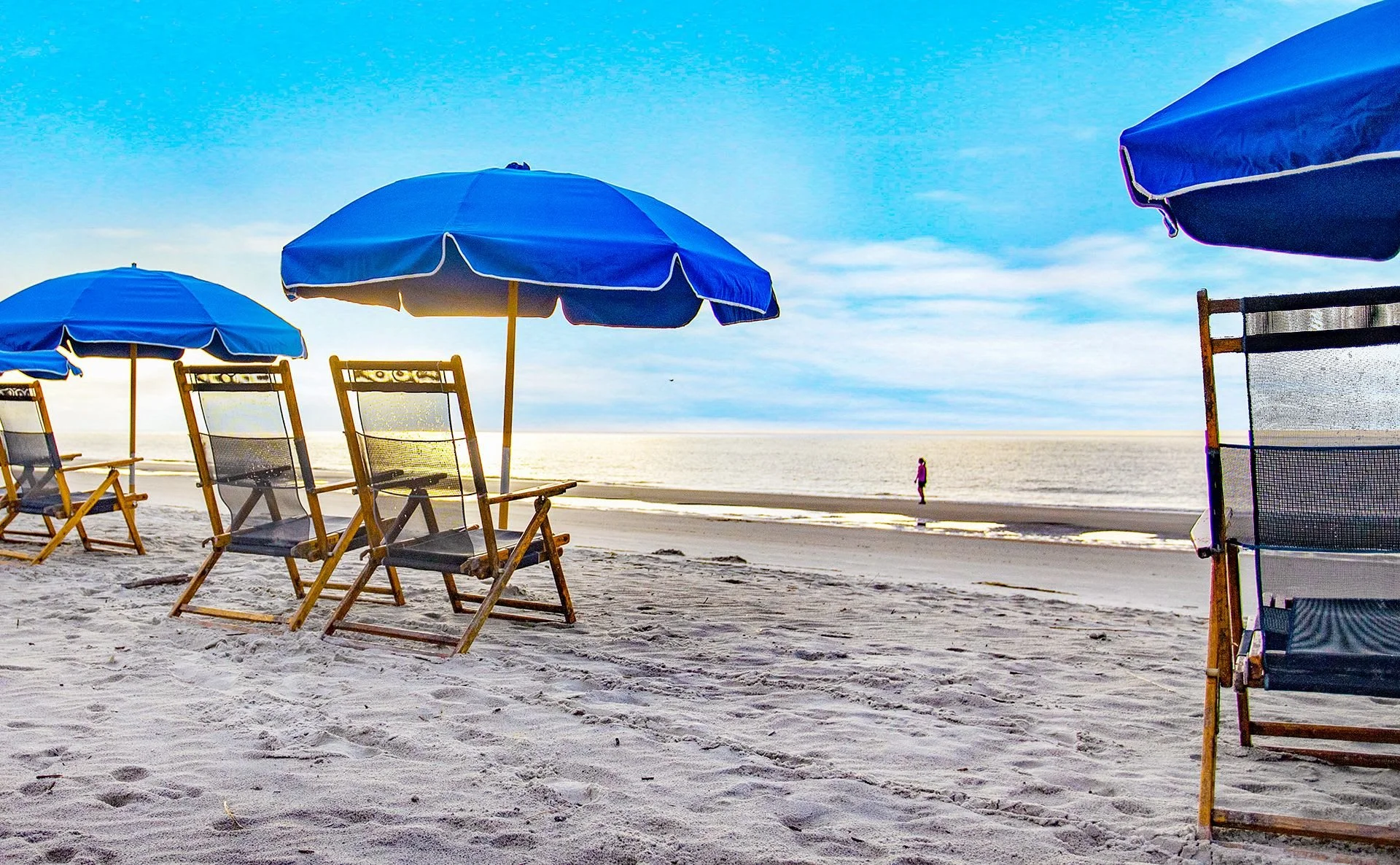 Beach scene with blue umbrellas and lounge chairs on sandy shore, ocean in background, person walking along water's edge.