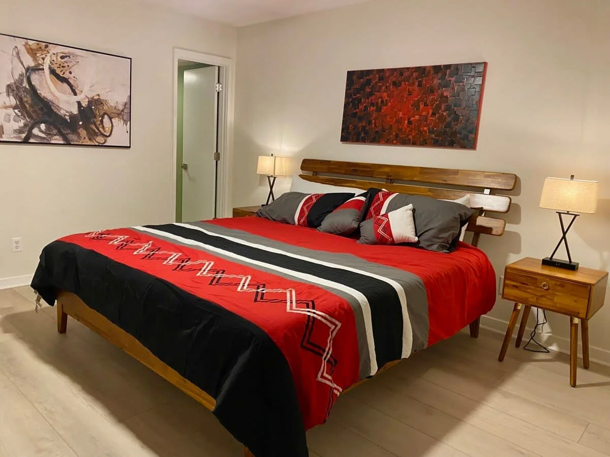 Modern bedroom with a wooden bed frame and red, black, and gray bedding, surrounded by abstract artwork on the walls and illuminated by bedside lamps on wooden nightstands.