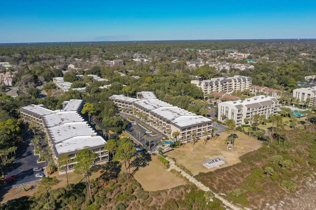 Aerial view of a resort complex surrounded by forested areas and greenery, featuring multiple buildings with flat roofs, parking lots, and well-maintained landscaping.