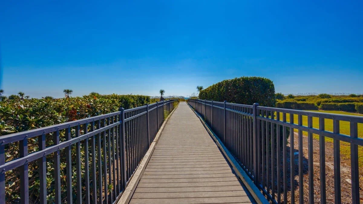 Wooden boardwalk with railings leading through greenery under a clear blue sky.