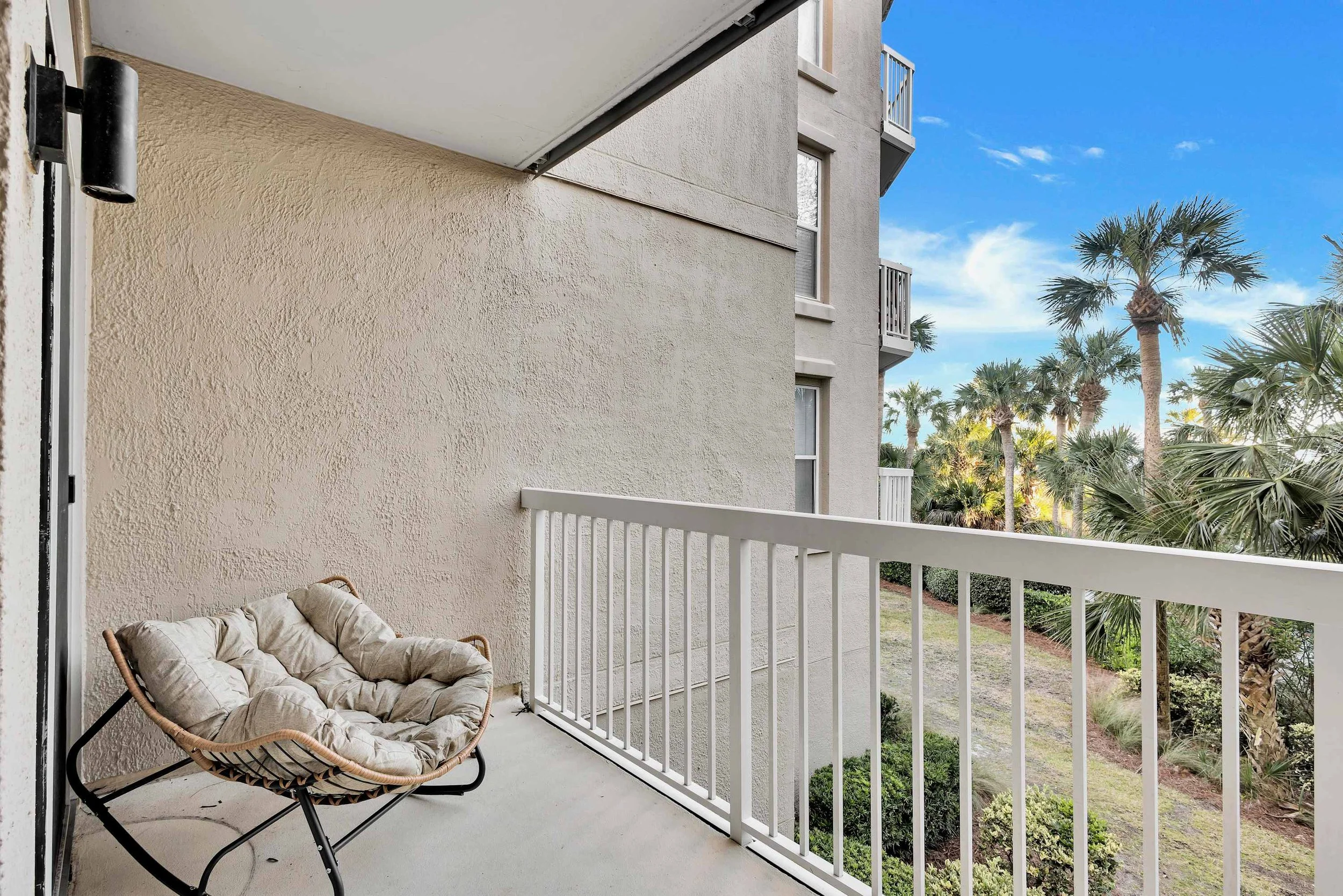 A balcony with a cushioned chair, white railing, stucco wall, and view of palm trees under a clear blue sky.