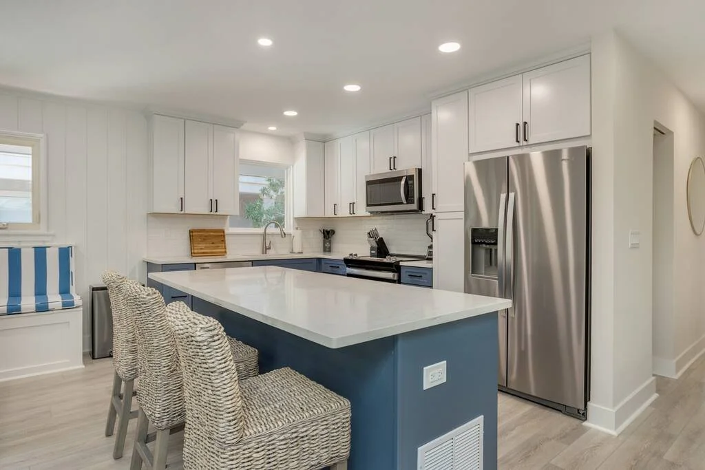 Modern kitchen with blue island, wicker bar stools, stainless steel appliances, white cabinets, and wooden floor.
