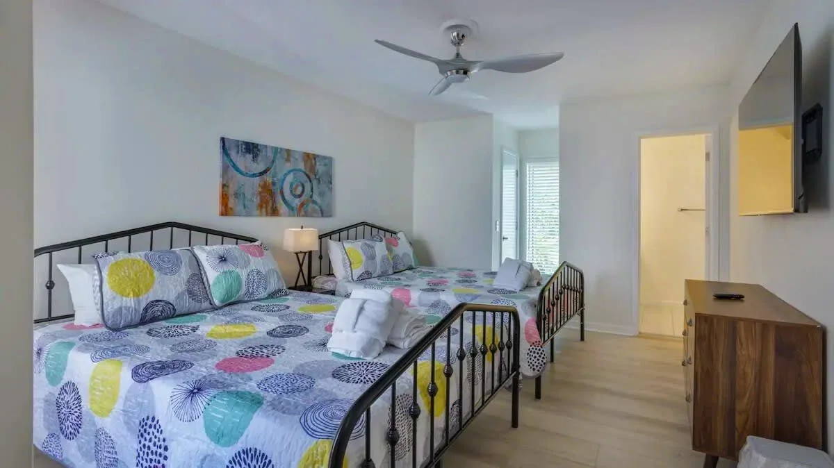 Modern bedroom with two metal-frame beds, colorful bedding, a ceiling fan, wall art, dresser, and a TV mounted on the wall.