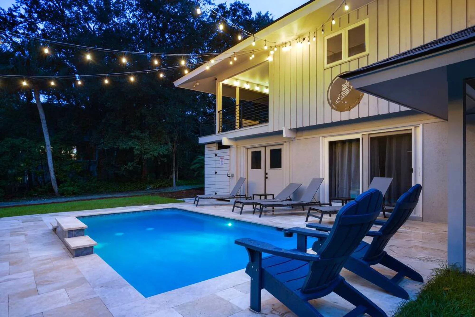 Backyard with a swimming pool, Adirondack chairs, lounge chairs, string lights, and a two-story house in the background at dusk.