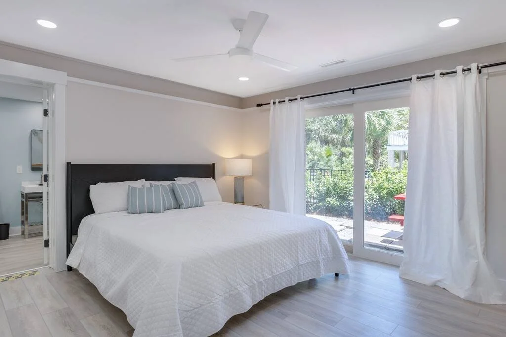 Modern bedroom with a white bed, pillows, and side lamp next to large sliding glass doors with white curtains leading to an outdoor patio.