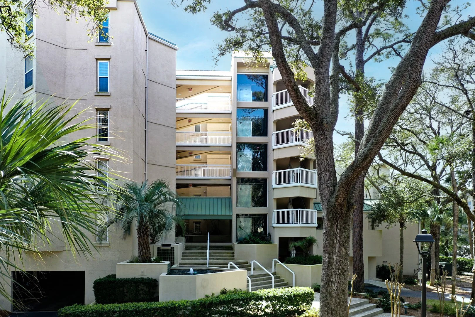Modern beige apartment building with balconies, surrounded by trees and greenery, featuring a small fountain in front.
