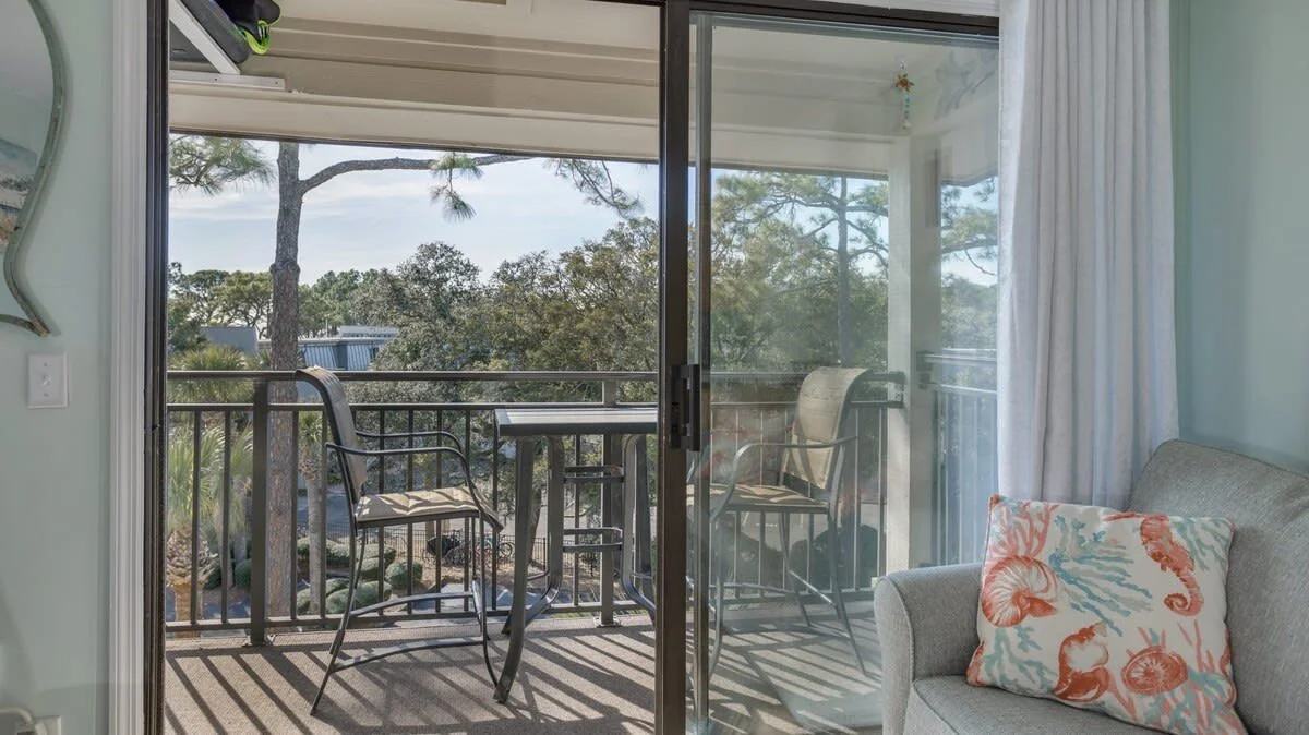 Living room with sliding glass door leading to a balcony with outdoor seating, overlooking trees.