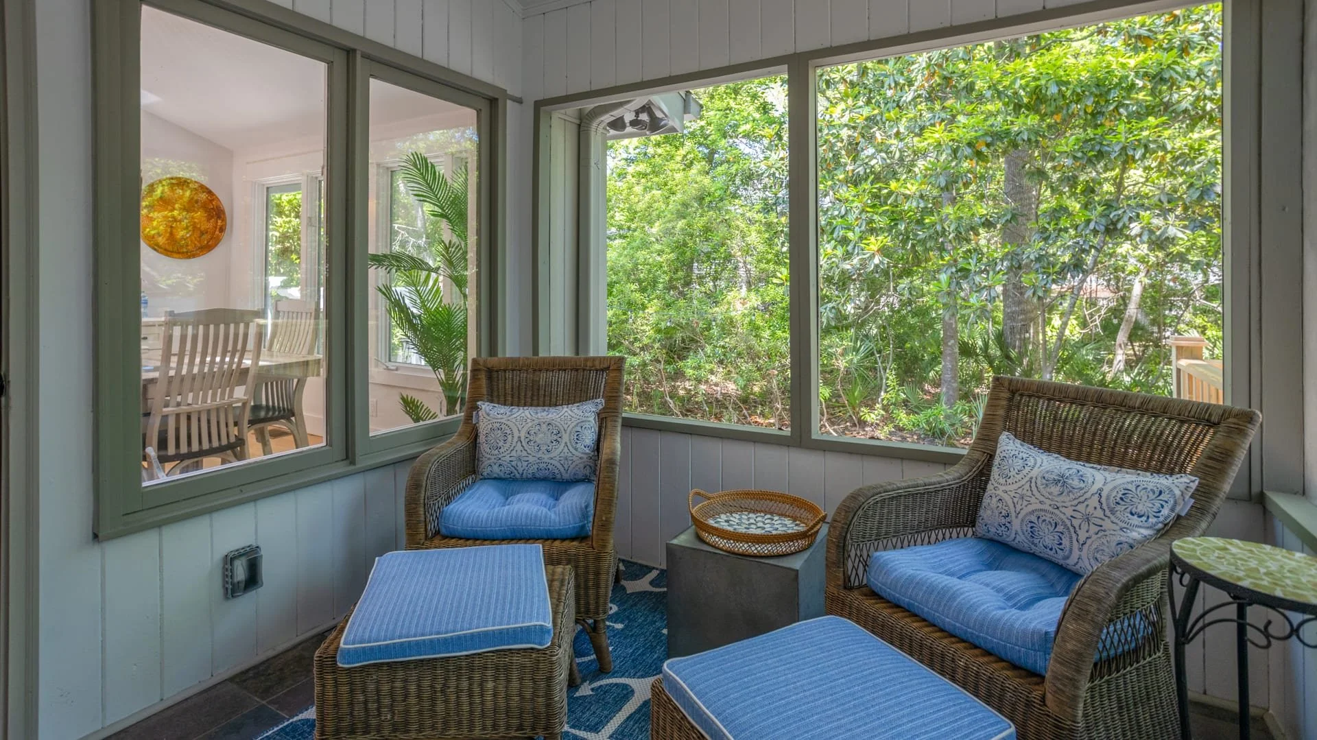Screened porch with wicker furniture, including two chairs with blue cushions and patterned pillows, a small round table, and a view of a garden through large windows. Inside, a dining area with wooden chairs is visible.