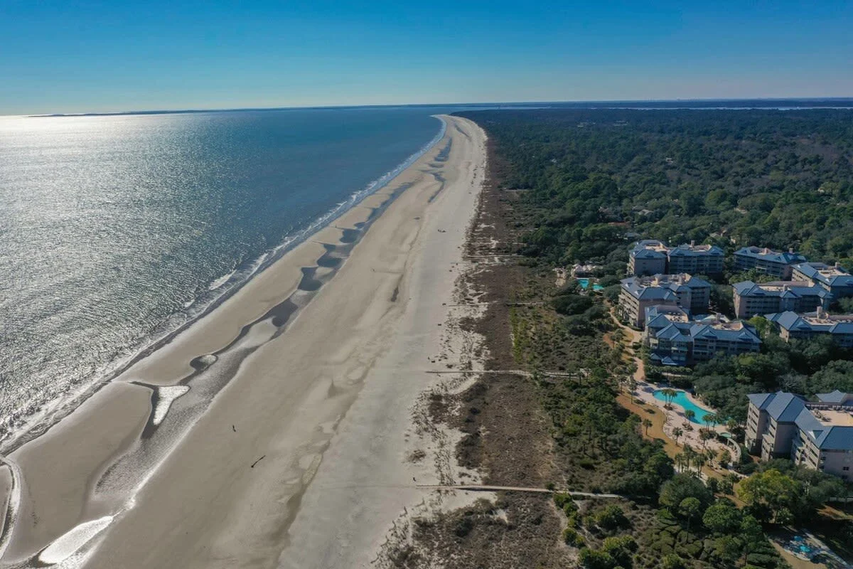 Aerial view of a long, sandy beach with ocean waves on one side and greenery with buildings on the other, under a clear blue sky.