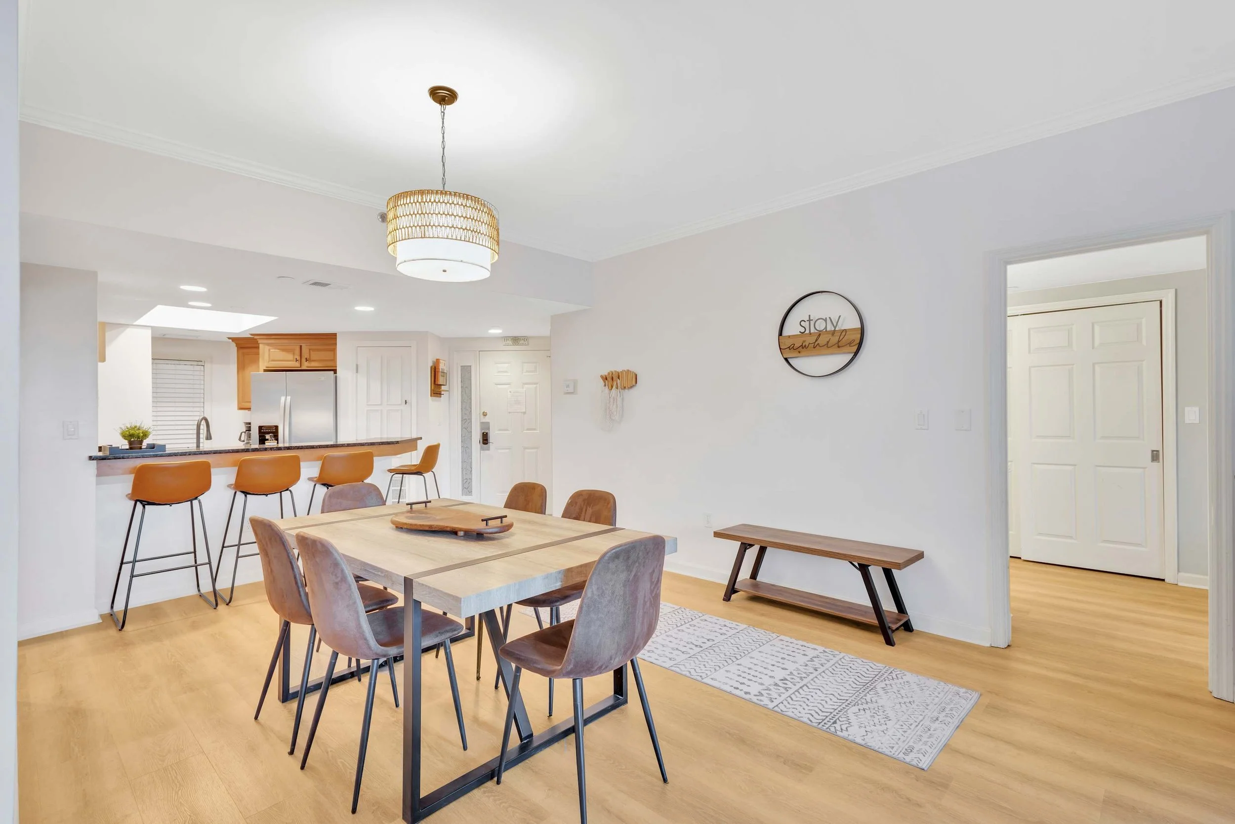 Modern dining room and kitchen area with wooden furniture, pendant light, bench, wall decor, and a counter with bar stools. Open layout with light wood flooring and white walls.