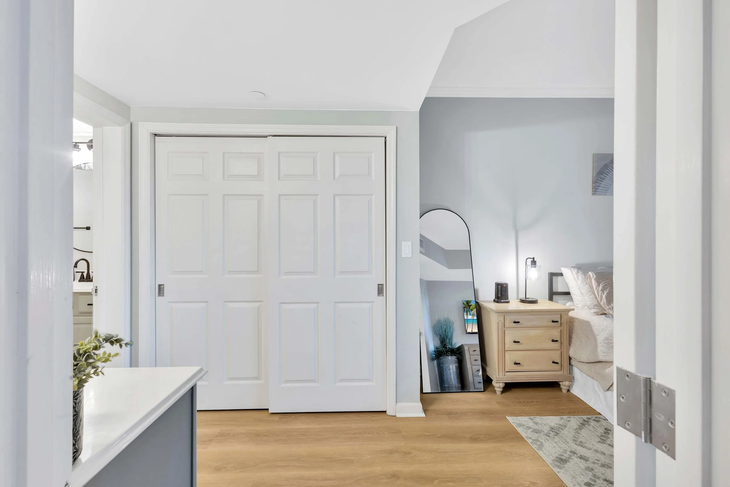 Bedroom with wooden floor, white closet doors, a nightstand with a lamp, and a leaning full-length mirror.