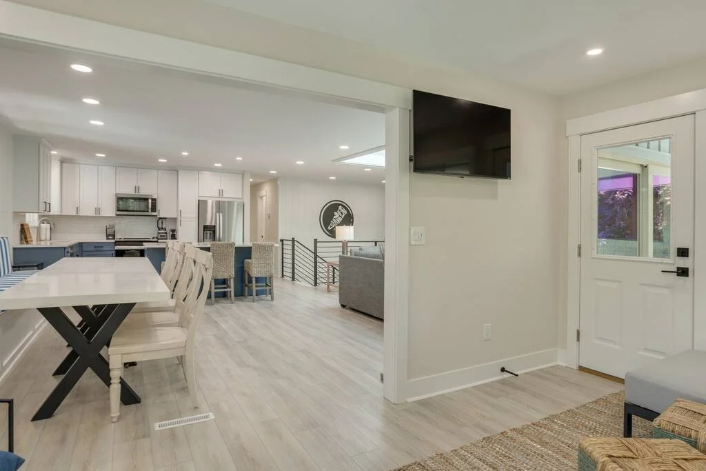 Modern open-concept kitchen and dining area with white cabinets, large dining table, overhead lighting, and a visible television and door on the right.