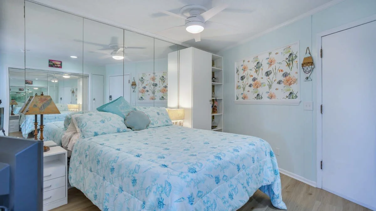 Bedroom with blue floral bedding, mirrored closet doors, white ceiling fan, floral wall art, and light wood floor.