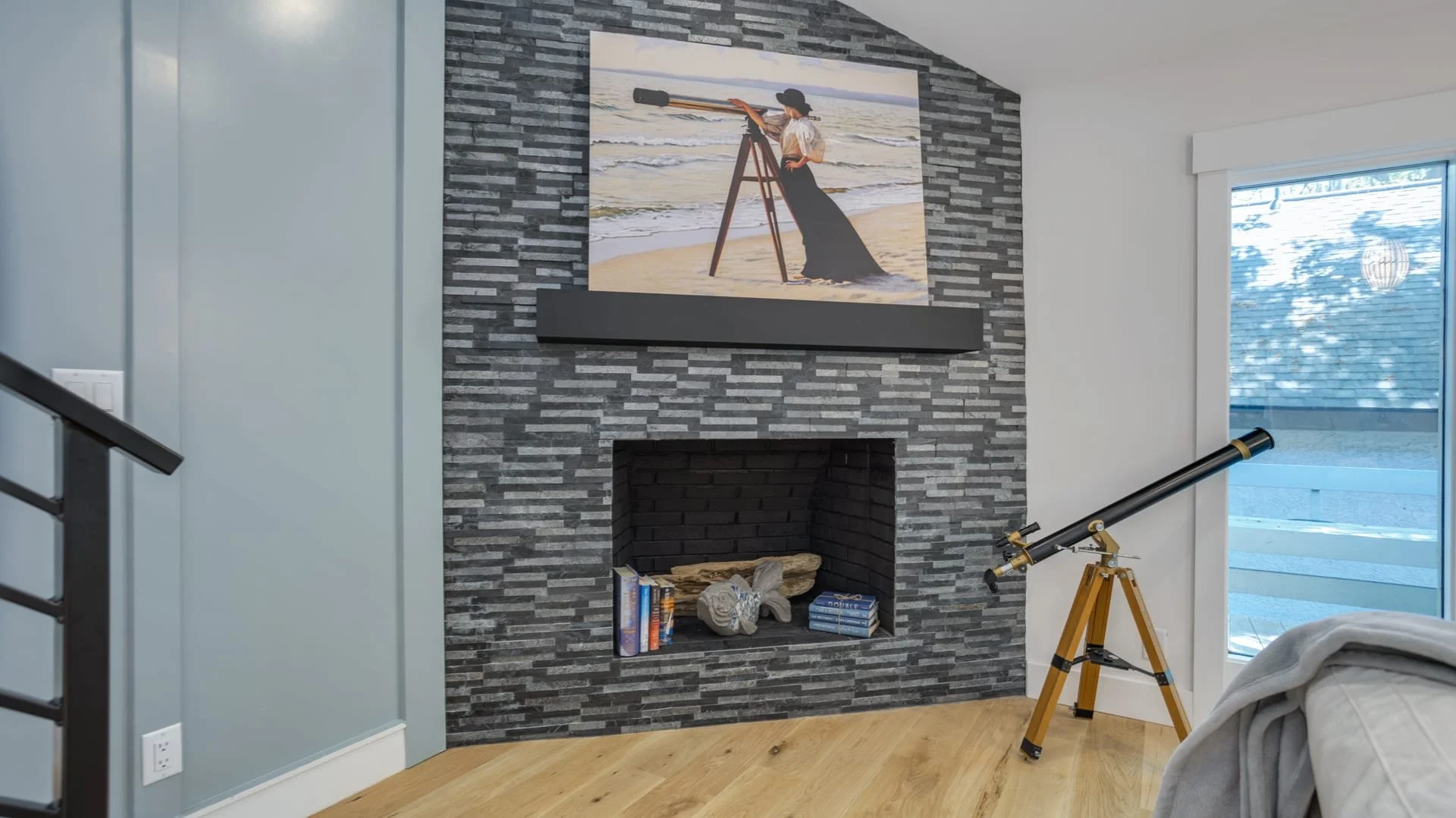Interior living room featuring a modern stone fireplace, an artwork of a person with a telescope on the beach, wooden flooring, and a telescope on a tripod next to a window.