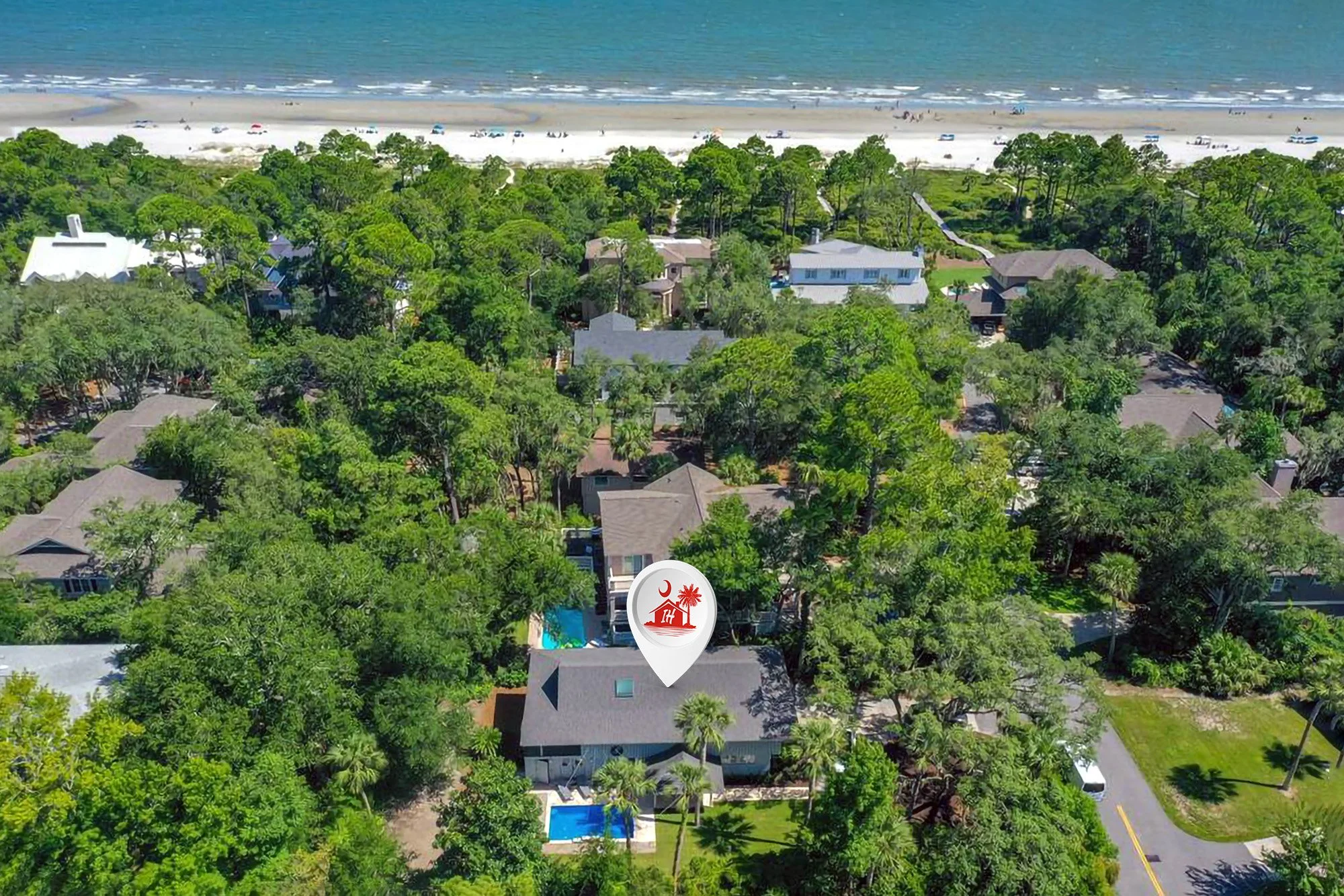 Aerial view of a coastal residential area with houses surrounded by lush green trees, a white sandy beach, and the ocean in the background. A location marker is centered over one of the houses.