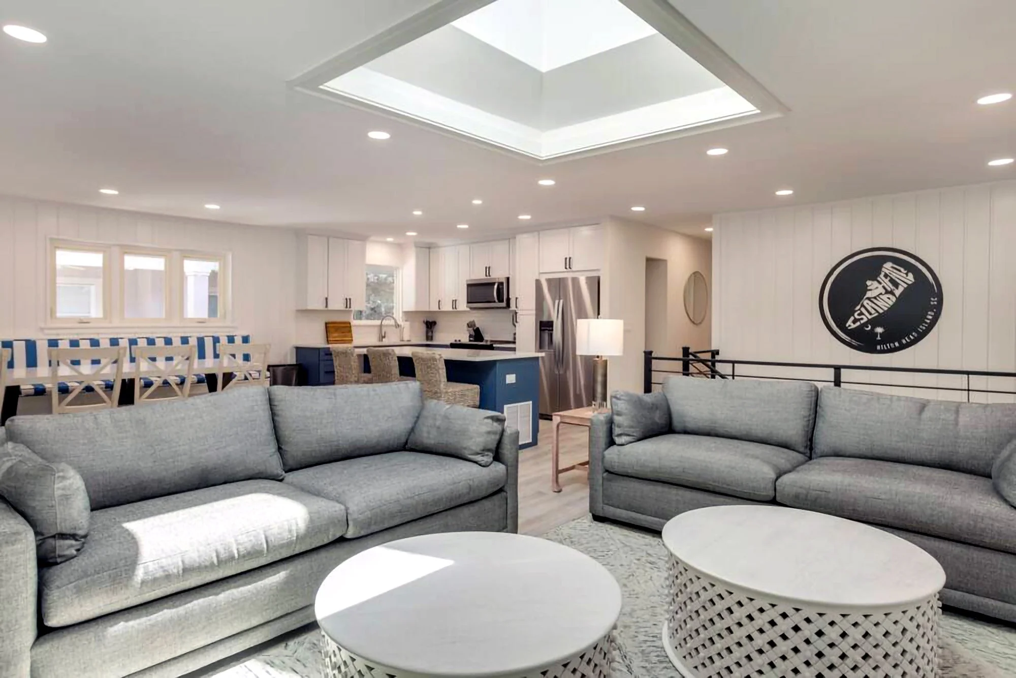 Modern living room with gray sofas, round coffee tables, dining area, open kitchen with island, and skylight ceiling.