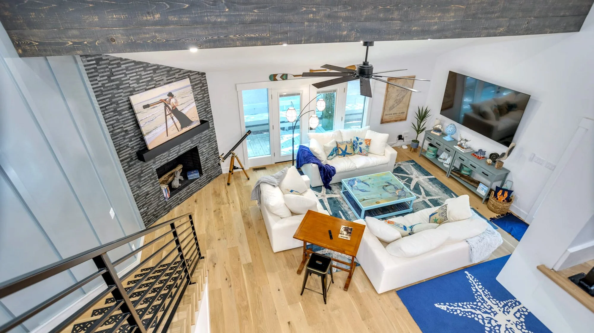 Modern living room with white sofas, wooden floor, and large TV. Features a fireplace with a painting above, a blue coffee table, and a telescope near glass doors. Decor includes a ceiling fan, indoor plants, and a nautical-themed rug.