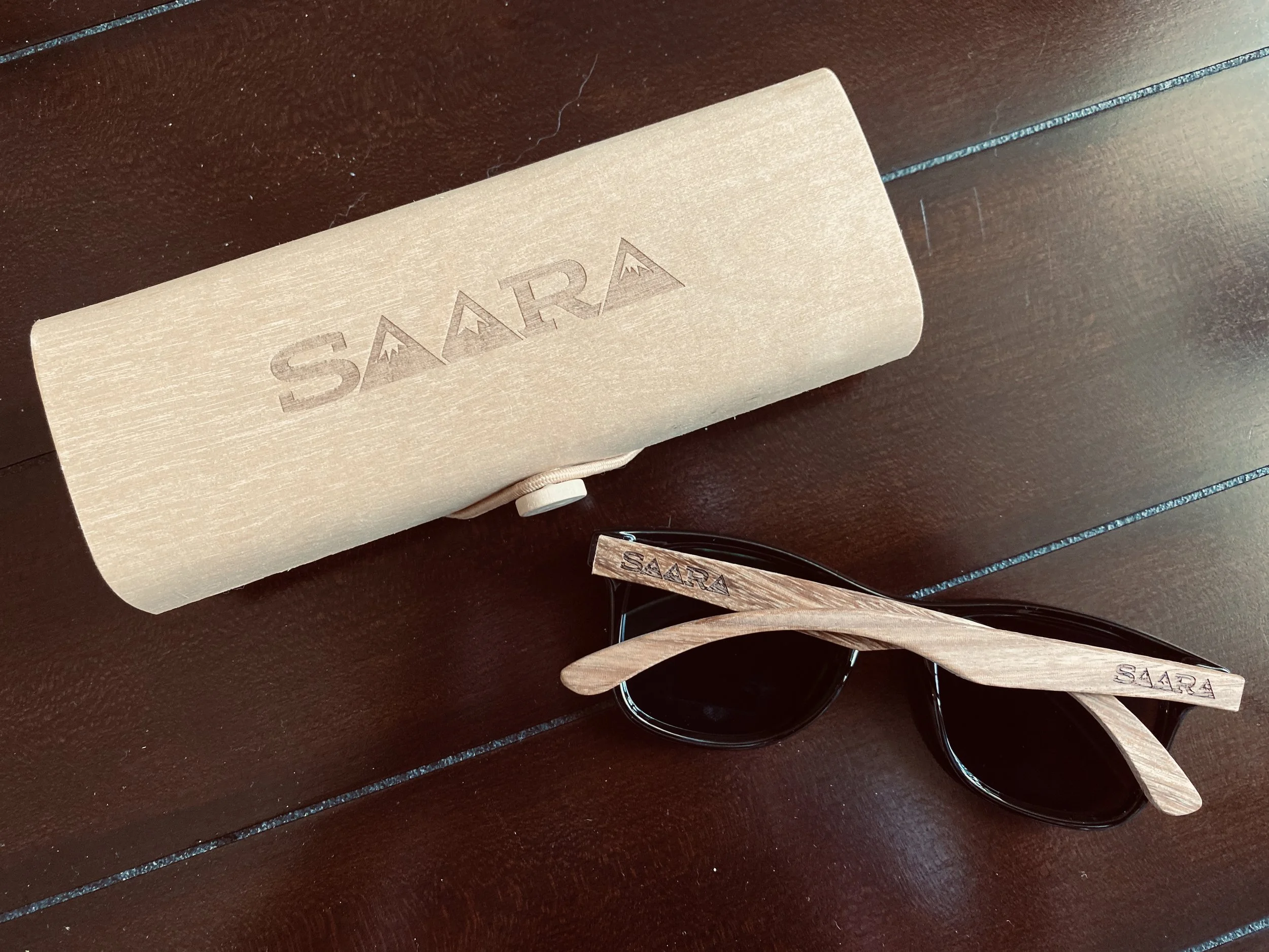 Wooden sunglasses with black lenses and a wooden case engraved with the word "SAARA" on a dark wooden surface.