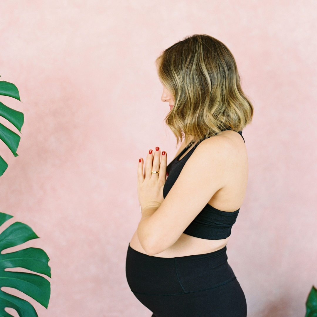 Pregnant woman in yoga pose with hands in prayer position, wearing black workout attire, standing against a pink background with green leaves nearby.