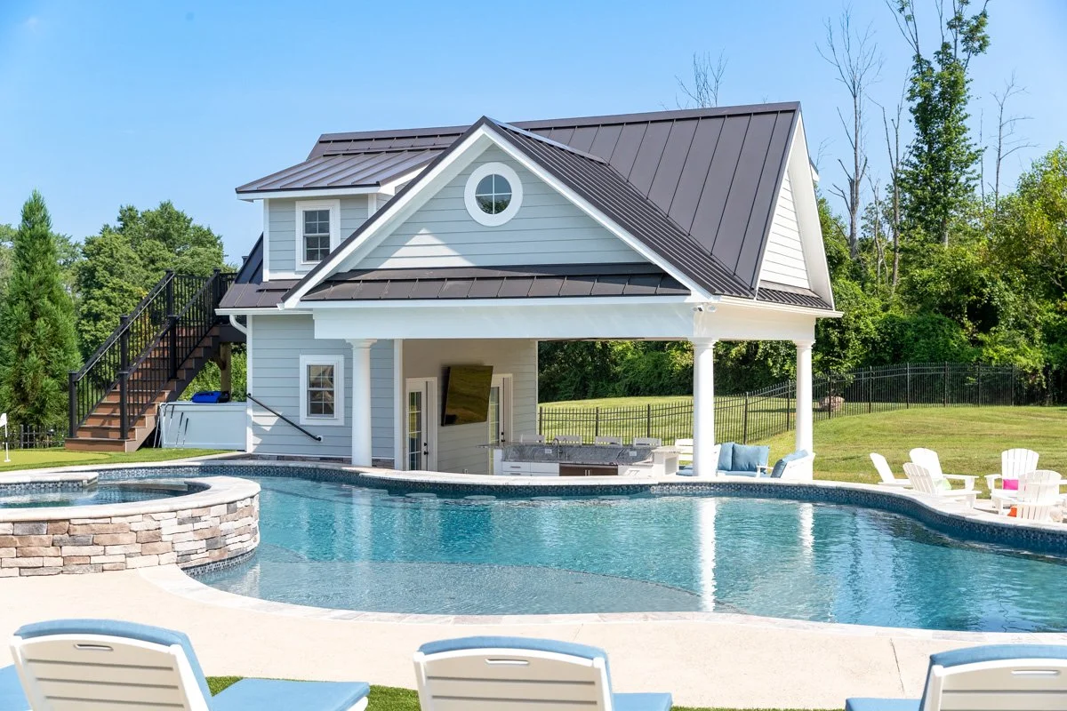 A backyard with a swimming pool and a white house with a metal roof, surrounded by green trees and lawn.